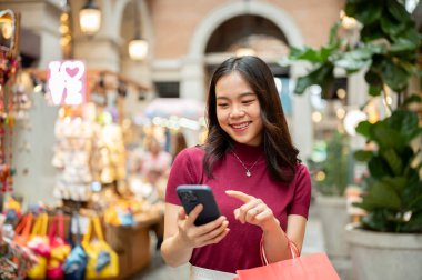 Alışveriş çantalı Asyalı bir kadın markette ya da alışveriş merkezinde hediyelik eşya arabasının yanında dururken telefona bakıyor. Ödeme günü tanıtımı, seyahat gezisi, turistik yerler..