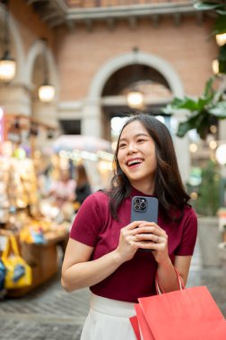 Alışveriş çantalı Asyalı bir kadın iki eliyle telefonu tutarken kapalı markette ya da alışveriş merkezinde dikiliyordu. Ödeme günü tanıtımı, seyahat gezisi, turistik yerler..