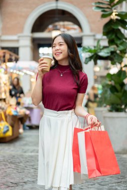 Asyalı kadın müşteri, alışveriş torbalarını kahve fincanıyla tutarken kapalı bir markette ya da alışveriş merkezinde yürürken. Ödeme günü tanıtımı, seyahat gezisi, turistik yerler..