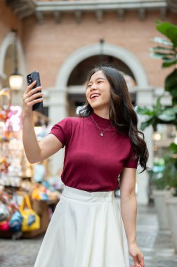 Asyalı bir kadın elinde telefonla gülerken ya da markette ya da alışveriş merkezinde yürürken. Ödeme günü tanıtımı, seyahat gezisi, turistik yerler..