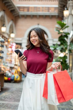Alışveriş çantası taşıyan gülümseyen Asyalı kadın telefona bakıyor kapalı markette ya da alışveriş merkezinde yürüyor. Ödeme günü tanıtımı, seyahat gezisi, turistik yerler..