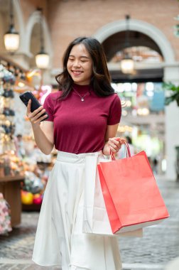 Alışveriş çantasıyla gülümseyen Asyalı kadın telefon kulübesine bakıyor ya da kapalı markette ya da alışveriş merkezinde yürüyor. Ödeme günü tanıtımı, seyahat gezisi, turistik yerler..