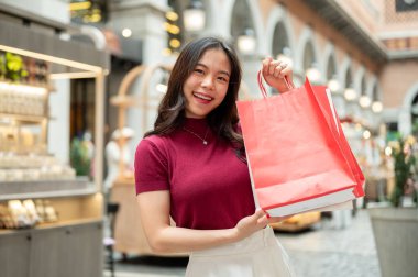 Kapalı bir markette ya da alışveriş merkezinde kırmızı alışveriş torbalarını tutan ve gösteren gülümseyen Asyalı kadın. Ödeme günü tanıtımı, seyahat gezisi, turistik yerler..
