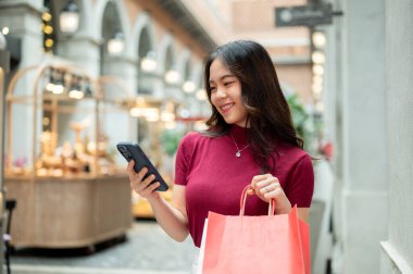 Asyalı bir kadın telefona bakıyor ve alışveriş çantalarını tutuyor. Kapalı markette ya da alışveriş merkezinde duruyor ya da yürüyor. Ödeme günü tanıtımı, seyahat gezisi, turistik yerler..