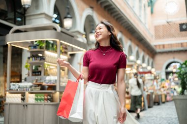 Gülümseyen Asyalı kadın alışveriş çantalarını tutarken yürürken ve etrafta dolaşırken veya alışveriş merkezinde. Ödeme günü tanıtımı, seyahat gezisi, turistik yerler..