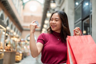 Elinde alışveriş çantası olan gülümseyen Asyalı kadın markette ya da alışveriş merkezinde dururken kredi kartına bakıyor. Ödeme Günü Tanıtımı, Seyahat Seyahati, Banka Reklamcılığı.