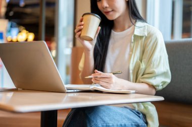 Kafedeki ahşap masada dizüstü bilgisayara bakarken kahve içen Asyalı bir kız öğrenci. Serbest Çalışan veya Çalışan, Modern Şehir Hayatı.