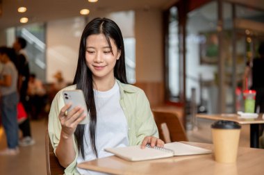 Mutlu Asyalı kız öğrenci telefona bakıyor ve kafedeki ahşap masada defter yanında kahve tutuyor. Serbest Çalışan veya Çalışan, Modern Şehir Hayatı.