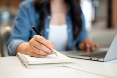 Not defterine kalem yazan kadın öğrenci kafedeki masada dizüstü bilgisayarın önünde oturuyor. Çalışmak ya da Çalışmak, Serbest Çalışmak, Şehir Hayatı.