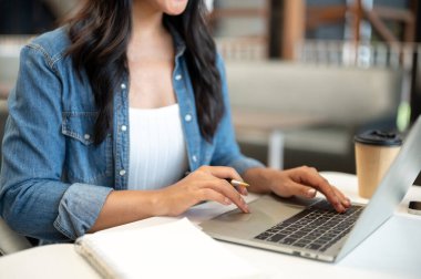Kalem tutan ve dizüstü bilgisayarla yazı yazan bir kız öğrencinin kafedeki masada otururken fotoğrafını çek. Çalışmak ya da Çalışmak, Serbest Çalışmak, Şehir Hayatı.