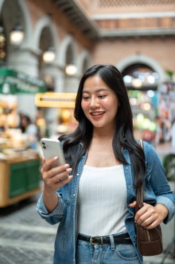 Asyalı bir kadın yürürken, alışveriş merkezinde ya da kapalı markette telefonuna bakıyor. Yalnız Seyahat, Turist Çekimleri, Unutulmaz Yolculuk.