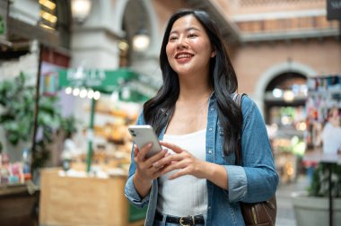 Asyalı kadın elinde telefonla yürürken ya da alışveriş merkezinde ya da kapalı markette dikilirken önüne bakıyor. Yalnız Seyahat, Turist Çekimleri, Unutulmaz Yolculuk.