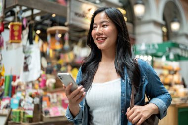 Asyalı bir kadın yürürken ya da alışveriş merkezinde ya da kapalı markette dururken telefon ve çanta askısı tutuyor. Yalnız Seyahat, Turist Çekimleri, Unutulmaz Yolculuk.
