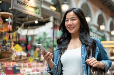 Asyalı bir kadın yürürken ya da alışveriş merkezinde ya da kapalı markette dururken telefon ve çanta askısı tutuyor. Yalnız Seyahat, Turist Çekimleri, Unutulmaz Yolculuk.