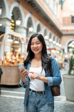 Asyalı bir kadın elinde telefonla dolaşırken ya da alışveriş merkezinde ya da kapalı markette dikilirken yüzünü çeviriyor. Yalnız Seyahat, Turist Çekimi, Unutulmaz Yolculuk.