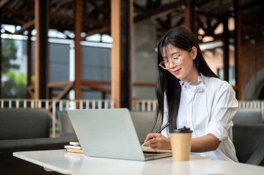 Gözlüklü Asyalı bir kız öğrenci kafeteryada dizüstü bilgisayarında kalem tutuyor. Serbest Çalışan veya Çalışan, Modern Şehir Hayatı.