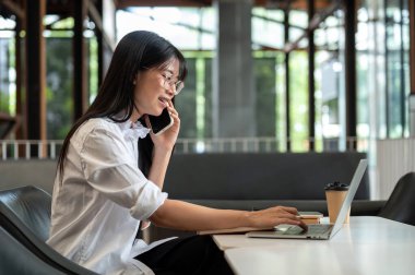 Gözlüklü Asyalı bir kız öğrenci telefonla konuşuyor ve kafedeki masada oturuyor. Serbest Çalışan veya Çalışan, Modern Şehir Hayatı.
