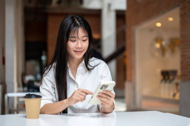 Bir binanın içinde masada otururken akıllı telefon kullanan ya da bakan Asyalı genç bir kadın. Modern Teknoloji, Boş Zaman, Şehir Günlük Hayatı.
