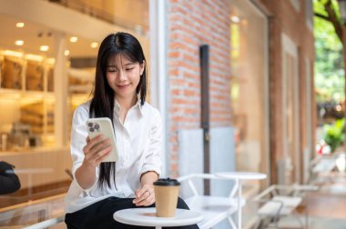 Asyalı genç bir kadın, tuğladan bir kafenin önündeki bankta otururken akıllı telefona bakıyor. Modern Teknoloji, Boş Zaman, Şehir Günlük Hayatı.
