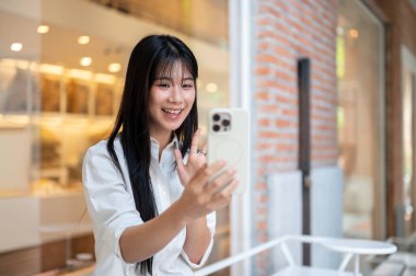 Asyalı genç bir kadın, duvardaki bir kafenin ya da mağazanın önünde otururken akıllı telefona el sallıyor. Modern Teknoloji, Boş Zaman, Şehir Günlük Hayatı.