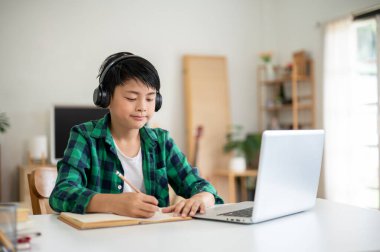 Asyalı ilkokul öğrencisi kulaklık takıp dizüstü bilgisayarın yanına kurşun kalemle yazı yazmış. Çevrimiçi Çalışma, Ödev yapma, İnternet Sörfü.