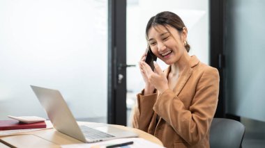 Asyalı işkadını ofis çalışanı ahşap bir masada dizüstü bilgisayarın önünde telefonla konuşuyor. Çalışma Saatleri, Şirket Masası İşi, Modern Şirket.