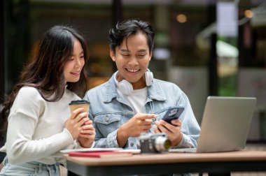 Telefona bakan genç Asyalı çift laptoplarını kafenin önündeki açık masada oturuyorlar. Randevu aktivitesi, Şehir Hayatı, Serinlemek Açık Hava