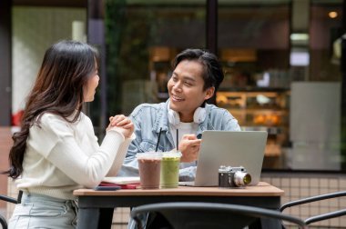 Kafenin önündeki masada içki fincanı ve dizüstü bilgisayarla konuşan genç Asyalı çift. Randevu aktivitesi, Şehir Hayatı, Serinlemek Açık Hava