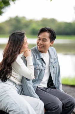 Genç Asyalı bir çift, parkta otururken gülümseyen bir kadına sarılıyor. Randevu aktivitesi, doğa iyileştirme, dışarıda dinlenme..