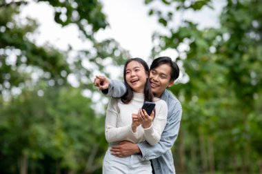 Genç Asyalı bir çift, parkta beklerken bir kadını işaret edip kucaklıyor. Randevu aktivitesi, doğa iyileştirme, dışarıda dinlenme..
