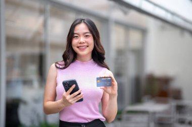 Genç Asyalı bir kadın bir kafenin önünde dururken elinde kredi kartı tutuyor. Banka reklamcılığı, Şehir Hayatı, Serinletici Açık Hava.