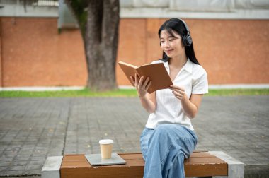 Genç Asyalı kız öğrenci kaldırım kenarındaki ahşap bankta otururken kulaklık takıyor ve kitap okuyor. Okul ya da üniversite, kampüs hayatı, boş zaman..