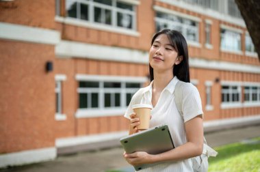 Laptop ve kahve fincanı tutan Asyalı genç bir kız öğrenci tuğla duvar ve bahçe boyunca yürüyor. Okul ya da üniversite, kampüs hayatı, boş zaman..