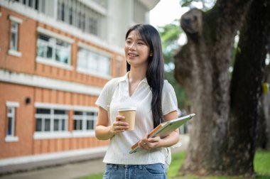 Dizüstü bilgisayar ve kahve fincanı tutan Asyalı genç kız bir binanın bahçesinde duruyor. Okul ya da üniversite, kampüs hayatı, boş zaman..