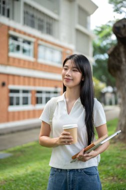 Genç Asyalı kız öğrenci dizüstü bilgisayarında kahve fincanıyla binanın bahçesinde duruyor. Okul ya da üniversite, kampüs hayatı, boş zaman..