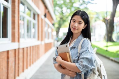 Sırt çantalı genç Asyalı kız öğrenci kaldırım kenarında kucak dolusu kitap ve dizüstü bilgisayar tutuyor. Okul ya da üniversite, kampüs hayatı, boş zaman..