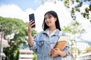 Bilgisayarı ve kitabı elinde tutan Asyalı bir kız öğrenci telefona bakıyor ya da dışarıda beklerken fotoğraf çekiyor. Okul ya da üniversite, kampüs hayatı, boş zaman..