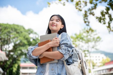 Güneşli bir günde açık havada dizüstü bilgisayar ve kitap tutan Asyalı bir kız öğrenci. Okul ya da üniversite, kampüs hayatı, boş zaman..