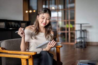 Tabletine bakıp gülümseyen Asyalı bir kadın kafede ya da kahvecide tahta bir sandalyede stilus kaldırıyor. Serinletici Açık Hava, Çalışma ya da Çalışma.