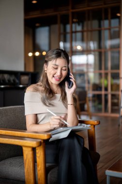 Bir telefon görüşmesi sırasında gülümseyen Asyalı kadın tabletini kafede ya da kahvecide tahta bir sandalyeye çiziyor. Serinletici Açık Hava, Çalışma ya da Çalışma.