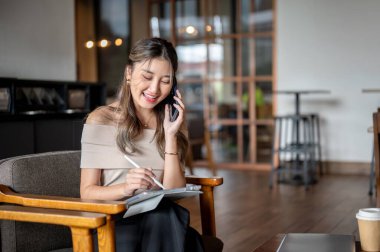 Telefon görüşmesi sırasında gülümseyen Asyalı kadın tabletine, kafede ya da kahvecide tahta bir sandalyeye yazı yazıyor. Serinletici Açık Hava, Çalışma ya da Çalışma.