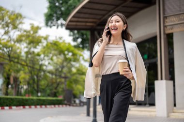 Uzun saçlı Asyalı kadın kahve fincanı tutuyor ve binanın önündeki kaldırımda yürürken telefonla gülümsüyor. Şehir Hayatı, Dışarıdaki Serinleme.