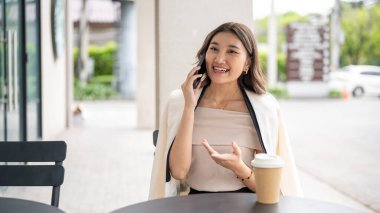 Uzun saçlı Asyalı kadın telefonda konuşurken elini açıklarken gülümsüyor kafedeki siyah masada kahve fincanı ile. Şehir Hayatı, Dışarıdaki Serinleme.