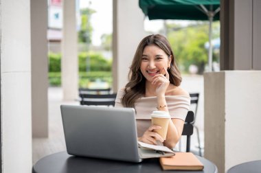 Kafe masasında dizüstü bilgisayarlı kahve fincanı tutan uzun saçlı Asyalı kadın, yanağına dokunarak gülümsüyor. Çalışıyorum ya da çalışıyorum, dışarıda takılıyorum..