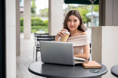 Uzun saçlı Asyalı kadın elinde laptop ve kitaplarla kafeterya masasında kahve fincanı tutarken kameraya bakıyor. Çalışıyorum ya da çalışıyorum, dışarıda takılıyorum..