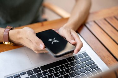 Chiang Mai, Tayland - 8 Mayıs 2025: Ahşap masadaki dizüstü bilgisayarda X uygulaması (Twitter 'da daha önce olduğu gibi) kullanarak akıllı telefonu tutan bir adama yaklaş.