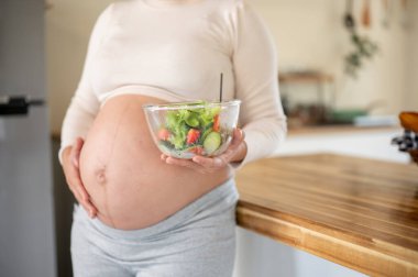 Hamile bir kadının elinde cam bir kase sebze salatası tutarken ve mutfakta karnına dokunurken yakın plan. Doğum öncesi bakım, Hamilelik Diyet yemeği.