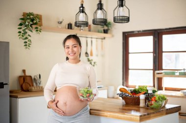 Hamile Asyalı kadın, elinde cam bir kase sebze salatası tutarken mutfakta çıplak karnına dokunuyor. Doğum öncesi bakım, Hamilelik Diyet yemeği.