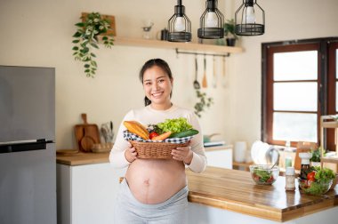 Hamile Asyalı kadın mutfakta dikilirken salata yapmak için elinde taze sebze sepeti ve ekmek tutuyor. Doğum öncesi bakım, Hamilelik Diyet yemeği.