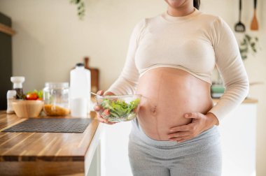 Hamile bir kadının elinde cam bir kase sebze salatası tutarken ve mutfakta karnına dokunurken. Doğum öncesi bakım, Hamilelik Diyet yemeği.